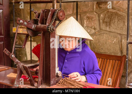 A Vietnamese woman wearing a traditional bamboo conical hat makes incense sticks using an old machine. Stock Photo
