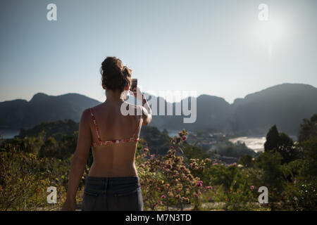 Tanned girl stands on a hill and takes a photo of the beautiful landscape. Back view of unrecognizable young woman. Tropical travel destination Stock Photo