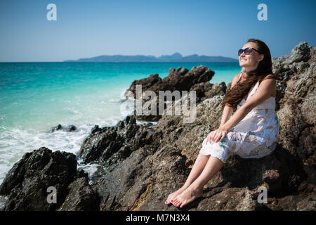 Young woman in dress sits on the rock on the coast of the ocean. Smiling girl is enjoying sunny summer day. Tropical tavel destination Stock Photo