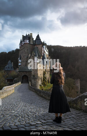 Young unrecognizable woman stands back on road leading to a beautiful castle. Fabulous medieval place. Trendy girl is traveling in Europe Stock Photo