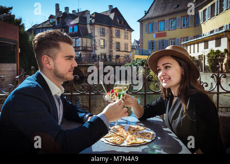 Young woman with boyfriend lunching in Alsace. Two people sits in local cafe with pizza and wine and holds wine glasses on terrace in romantic place Stock Photo