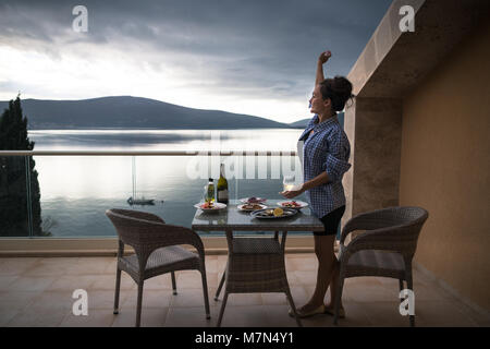 Young woman standing on balcony of apartment near table with food and drinks and waving to somebody. Smiling girl with glass of wine looking on sea Stock Photo