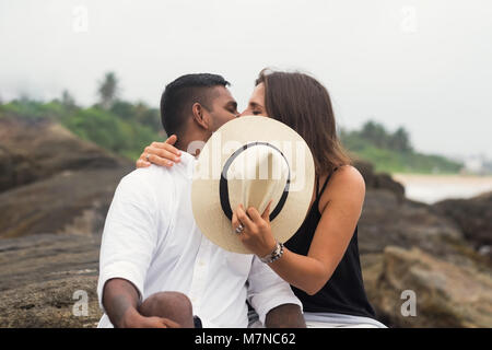 Young mixed race couple kissing sitting on stone. They close faces with hat. Stock Photo