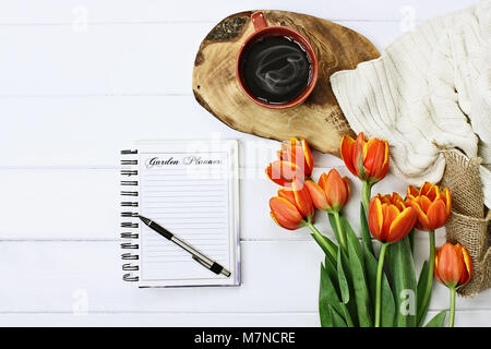 Overhead shot a garden journal notebook with pen, coffee and a bouquet of orange and yellow tulips over a wood table. Flat lay top view style. Stock Photo