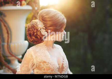 Blonde woman with elegant hairstyle wearing dress and white lace jacket, standing on garden terrace and leaning on balustrade decorated with large flo Stock Photo