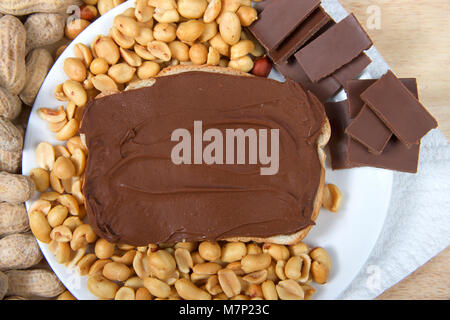 Chocolate peanutbutter spread on bread, open face sandwich on plate with pieces of chocolate, shelled peanuts and whole in shell peanuts on wood table Stock Photo