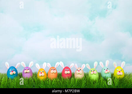 Colorful decorated Easter Eggs with bunny faces and ears lined up in a row on green grass, vintage sky background. Fun Easter concept. Stock Photo