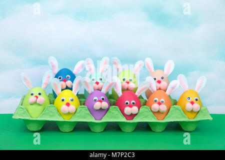 Colorful decorated Easter Eggs with bunny faces and ears sitting in an egg carton on green table, vintage sky background. Fun easter concept. Stock Photo