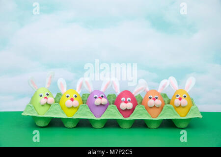 Colorful decorated Easter Eggs with bunny faces and ears sitting in an egg carton on green table, vintage sky background. Fun easter concept. Stock Photo