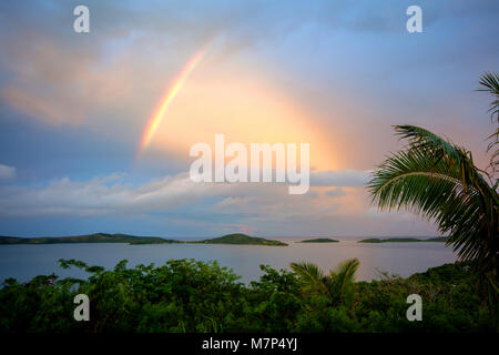 Beautiful landscape of Fiji island during high tide with palm trees, bushes in foreground and ocean with green hills and very unique rainbow in sky. Stock Photo