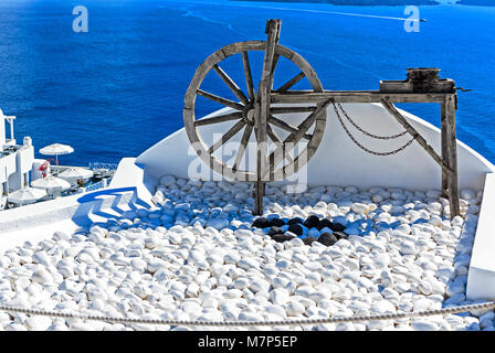 Ancient wooden wheel and white painted pebbles against blue Aegean Sea in Santorini (Oia), Greek Islands, Greece Stock Photo