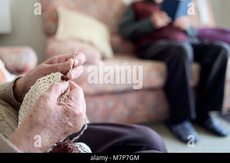 closeup of an old caucasian woman crocheting a piece with a beige thread and an old man reading a book sitting on a couch in the background Stock Photo