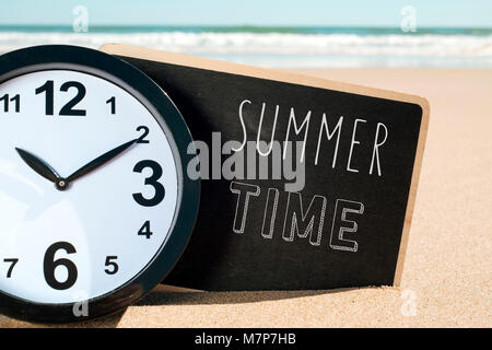 closeup of a clock and a signboard with the text summer time written in it, on the sand of a beach Stock Photo