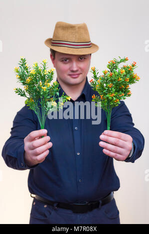 Portrait of young elegant man with blue shirt and summer hat holding artificial yellow and orange flowers in both hands. Stock Photo