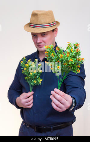 Portrait of young elegant man with blue shirt and summer hat holding artificial yellow and orange flowers in both hands. Stock Photo