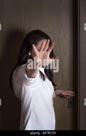 Caucasian young woman protecting herself from being seen by putting her hand in front of the camera, while standing at a closed wooden door. Stock Photo