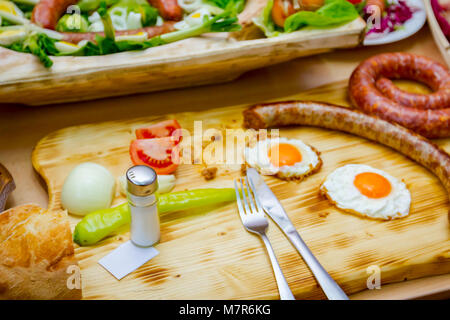 Fried eggs and sausage are placed on a wooden chopping board with cutlery, saltshaker. Stock Photo