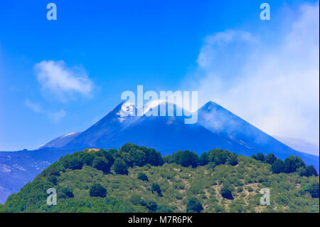 Picturesque view of Mt Etna in Sicily on a sunny day. Lush green volcanic landscape with majestic mountain shrouded in wispy clouds, against blue sky Stock Photo