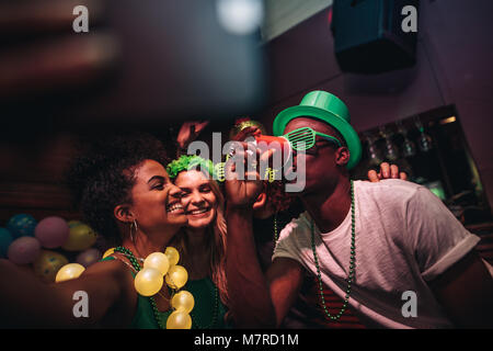 Man drinking a glass of beer with female friends smiling and taking selfie at bar. Young people celebrating St.Patricks day at night club and taking s Stock Photo