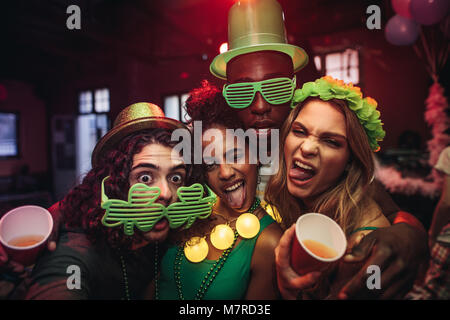 Crazy young friends enjoying a party at pub. Men and women in green clover party glasses and leprechaun hat at night club. People celebrating St.Patri Stock Photo
