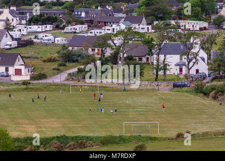 Aultbea, Scotland - June 9, 2012: Focus on football field with young players in middle of the village with white houses and cars. Green vegetation. Stock Photo