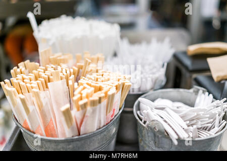 Many disposable wooden chopsticks in paper wrappers in bowl in Asian Japanese restaurant fast food casual cafe by plastic cutlery, forks, spoons Stock Photo