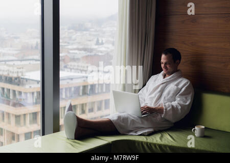 Young smiling businessman working on laptop computer wearing white bath robe sitting near window with cup of coffee looking at sunrise city view. Motivation concept Stock Photo