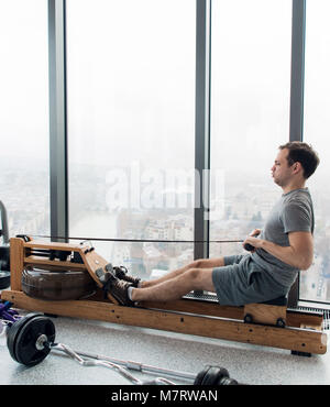 Rowing with power. Side view of young man in sportswear doing rowing in front of window at gym. Stock Photo