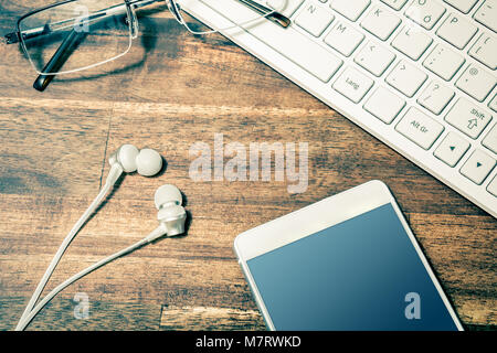 Closeup Of A White Mobile Next To In-Ear Headphones, Glasses And White Keyboard On Wooden Table Stock Photo