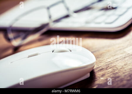 Macro Of A White Computer Mouse Lying Next To Glasses And White Keyboard On Wooden Table Stock Photo