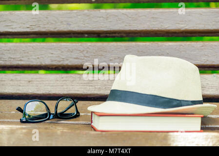 straw hat, glasses and a thick book on a wooden bench in the park Stock Photo