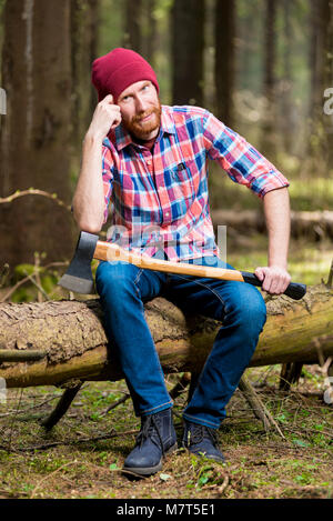 thoughtful lumberjack in a hat and shirt with an ax sits on a log in the woods Stock Photo