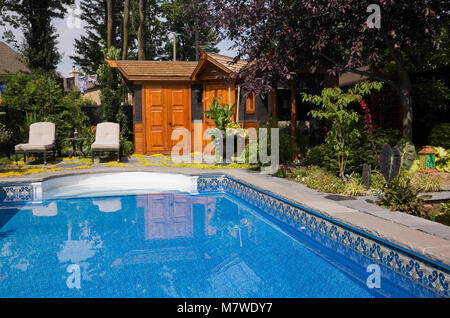 Swimming pool and long chairs on patio stones next to a storage shed in a landscaped residential backyard in summer. Stock Photo