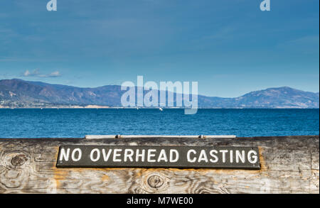 wooden sign on tree trunk barrier saying No Overhead Casting at the Pacific Ocean in Santa Barbara, California, USA with mountains in the distance Stock Photo