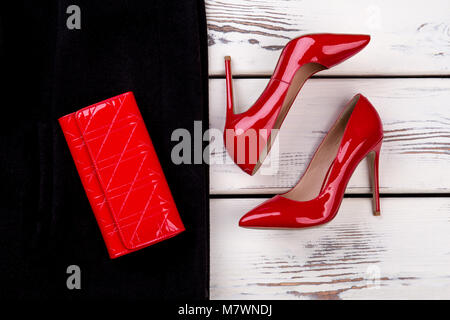 Close up women's red shoes and wallet. Leather red wallet on black clothes. Bright beige white wooden desks surface background. Stock Photo
