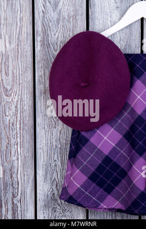 Purple beret hat and checkered skirt. Flat lay, top view. Dark wooden desk surface background. Stock Photo