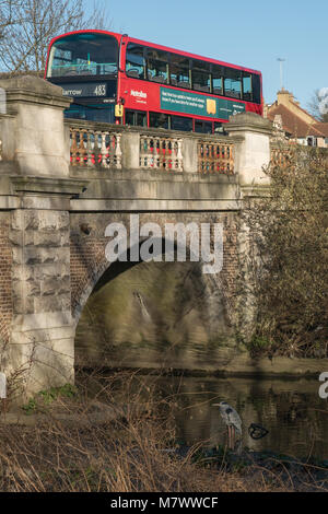 A crane (bird) on the river Brent as a red bus passes over a bridge near Hanwell in west London. Photo date: Sunday, February 25, 2018. Photo: Alamy Stock Photo