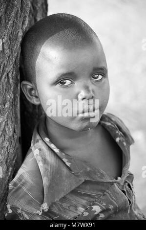 Monochrome portrait of a young boy leaning against a tree trunk in his local village. There is still much curiosity surrounding foreigners in Africa Stock Photo