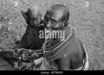 Monochrome portrait of an unidentified mother and child, wearing traditional ornamental necklace of red beads, sits outside hut in local village. Stock Photo