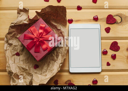 Gift in a beautiful red box with ribbon. The tablet with an empty screen is on the wooden surface next to the gift. An event, a holiday. Stock Photo