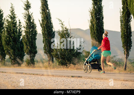 Running mother with child in baby stroller enjoying sunset and mountains landscape. Jogging or power walking supermom, active family at sunset. Beauti Stock Photo