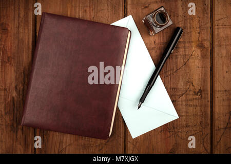 An overhead photo of a leather bound journal, a blue envelope, an ink well and pen, shot from above on a dark rustic background with a place for text Stock Photo