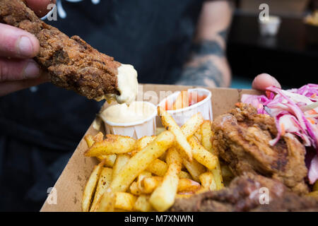 Fried chicken and chips with coleslaw, dipped in mayonnaise with ketchup by man with tattoos.  Hipster street food. Stock Photo