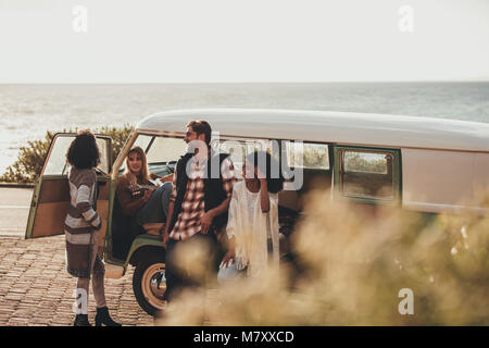 Friends hanging out together by a van. Group of man and women standing by an old minivan on roadside, taking rest during the road trip. Stock Photo