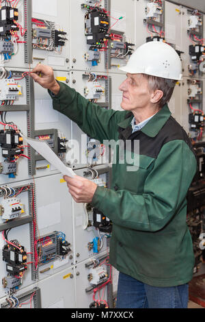 Senior electrician with a screwdriver and wiring diagram in his hand standing near an an electric shield Stock Photo