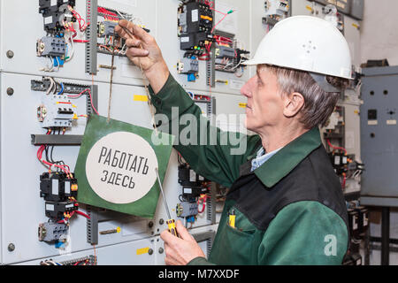 Senior electrician with a screwdriver sets a sign at an electric shield, nameplate with inscription in Russian: Work here Stock Photo