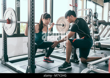 Woman doing squats with barbell. The personal trainer helping her flexing muscles in Smith machine in gym. Stock Photo