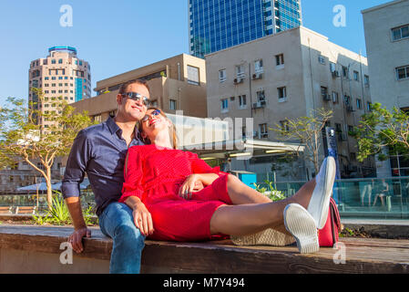 Young loving couple in bright clothes and sunglasse ssitting on the bench in the city area. Embracing, laughing ,relaxing. Happiness, lifestyle and to Stock Photo
