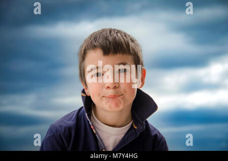 Little Boy is standing in front of the camera and making funny faces. Blurred dramatic sky as background. Stock Photo