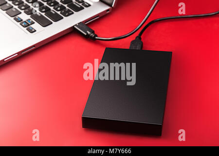 An external hard drive connected to the laptop with a usb cable on a red background. Portable storage technologies Stock Photo